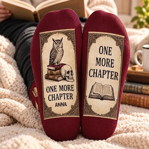 May include: A pair of deep red socks featuring a rectangular graphic on the soles. The left sole displays an owl perched on a stack of books next to a skull with the text ONE MORE CHAPTER and ANNA. The right sole shows an open book with the text ONE MORE CHAPTER.