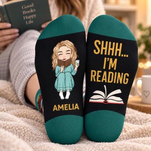 May include: A pair of black and dark green socks. The left sock features a cartoon illustration of a person with blonde hair reading a book and holding a cup, with the name AMELIA printed below. The right sock displays the text SHHH... I'M READING above an open book graphic.
