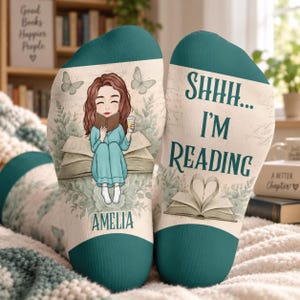 Op de afbeelding: Een paar blauwgroene en crèmekleurige sokken met een cartoonillustratie van een lezend persoon, vlinders en de tekst "AMELIA" op de ene sok, en "SHHH... I'M READING" met een open boek op de andere sok.