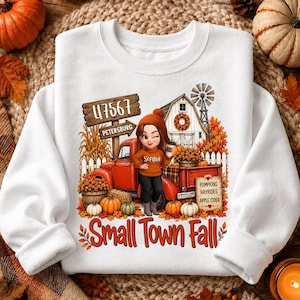May include: A white long-sleeve sweatshirt featuring a graphic print of a cartoon character in an orange sweater standing by a red truck. The design includes a barn, pumpkins, autumn leaves, and text reading 47567 PETERSBURG, PUMPKINS HAYRIDES APPLE CIDER, and Small Town Fall.