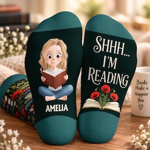 Op de afbeelding: Een paar donkergroen-blauwe sokken met een cartoonillustratie van een persoon met blond haar die een boek leest, de naam AMELIA en de tekst SHHH... I'M READING boven een open boek met rode klaprozen die uit de pagina's komen.