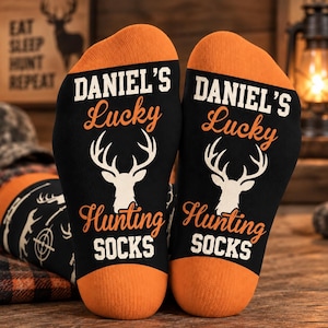 May include: A pair of black socks with orange heels and toes, featuring a white deer head graphic and the text DANIEL'S Lucky Hunting SOCKS printed in white and orange. The socks have a pattern of deer silhouettes and target symbols.