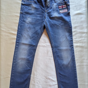 May include: Blue denim jeans featuring a faded wash, a button and zipper fly, and two rectangular patches on the upper right thigh. The top patch displays the Union Jack flag, while the bottom patch features the United States flag design.