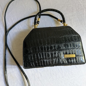 Puede incluir: Un bolso de mano negro con textura brillante de imitación cocodrilo, que presenta una forma trapezoidal estructurada, un asa superior y una correa larga para el hombro. Una placa metálica dorada con el texto BULAGGI está fijada en la parte frontal.
