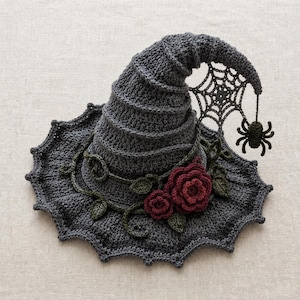 Könnte beinhalten: Ein dunkelgrauer, gehäkelter Hexenhut mit spitzer, spiralförmiger Krone und breiter, gewellter Krempe. Der Hut ist mit dunkelroten gehäkelten Rosen, grünen Blättern und einem hängenden gehäkelten Spinnennetz mit kleiner schwarzer Spinne verziert.
