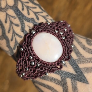 May include: A macrame bracelet featuring a large, smooth, oval-shaped white stone set in dark brown cord. The intricate knotted design is accented with small, round, metallic silver beads embedded throughout the woven pattern.