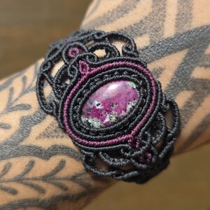 May include: A macrame bracelet featuring a central oval-shaped stone with mottled magenta, black, and white coloring. The stone is encased in a dark purple cord border, surrounded by an intricate black cord lattice pattern with decorative loops.