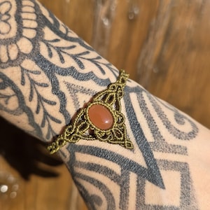 Puede incluir: Una pulsera de macramé con un cordón verde oliva tejido en un patrón decorativo con una piedra naranja lisa de forma ovalada en el centro. La pulsera está construida con técnicas de anudado intrincadas que forman un marco triangular.