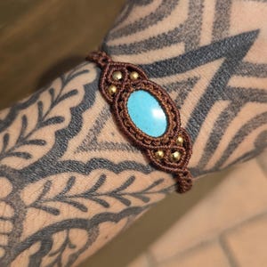 Könnte beinhalten: Ein braunes Makramee-Armband mit einem zentralen ovalen Türkisstein, der von kleinen metallisch goldenen Perlen umgeben ist. Das Armband besteht aus komplexen geknoteten Kordelmustern, die den leuchtend blauen Stein einrahmen und ein detailliertes Design bilden.