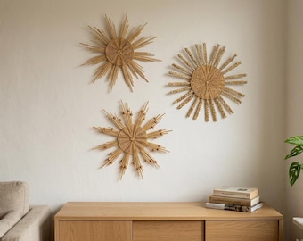 May include: Three circular wall decorations made of woven light brown natural fibers. Each piece features a central disc with radiating spokes of varying lengths and textures, creating a sunburst pattern. The designs are mounted on a plain wall.