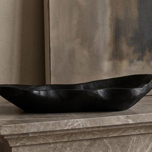 May include: An elongated, matte black decorative bowl with an organic, irregular, and wavy rim. The bowl features a deep, carved-out center and a smooth, dark finish, presenting a sculptural form that sits horizontally with a low profile.