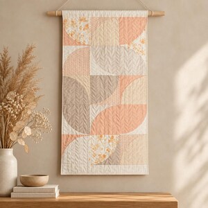 Könnte beinhalten: Ein rechteckiger, gesteppter Wandbehang mit einem geometrischen Muster aus Halb- und Viertelkreisen. Der Stoff zeigt eine Palette aus Pfirsich-, Creme- und Beigetönen mit verschiedenen Texturen, darunter einfarbige Stoffe, Fischgrätmuster und florale Drucke.