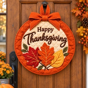 Puede incluir: Un tapiz redondo de tela acolchada con el texto bordado Happy Thanksgiving en marrón oscuro. El diseño incluye un gran lazo de tela naranja en la parte superior y tres hojas de fieltro en rojo, naranja y amarillo en el centro inferior.