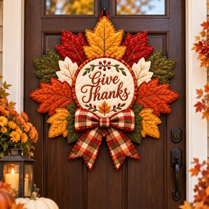 以下が含まれることがあります： Give Thanksという文字が中央の円形パネルに刺繍されたキルティング生地のリース。赤、オレンジ、黄色、緑の秋の葉が重なり合っており、リースの下部にはチェック柄の布製リボンが取り付けられています。