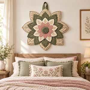 Puede incluir: Un tapiz acolchado en forma de estrella con pétalos de tela superpuestos en tonos verde salvia, crema y rosa empolvado. El diseño incluye costuras intrincadas, patrones florales y un motivo circular central, suspendido por una presilla de tela.