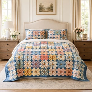 May include: A bed quilt and two matching pillow shams featuring a repeating floral patchwork pattern in shades of blue, yellow, pink, and white. The quilt has a blue border and a quilted texture, covering a bed with a light-colored headboard.