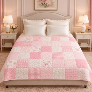 Op de afbeelding: Een patchwork sprei met een herhalend patroon van lichtroze en witte vierkanten. Het ontwerp wisselt af tussen effen roze, roze-witte vichy-ruitjes en witte stof met een verfijnde roze bloemenprint over het gehele oppervlak van de deken.