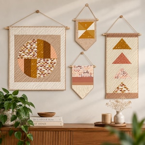 Peut inclure: Quatre tentures murales matelassées de tailles et formes variées, présentant des motifs géométriques dans des tons crème, beige, terre cuite et jaune moutarde. Chaque pièce est suspendue à une tige en bois avec une cordelette.
