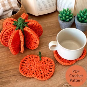 Può includere: Un set di sottobicchieri all'uncinetto arancioni a forma di zucca con steli verdi, incluso un supporto impilabile. Un sottobicchiere è posto sotto una tazza in ceramica bianca. Un cerchio arancione riporta il testo PDF Crochet Pattern.