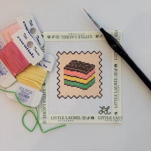 Puede incluir: Un lienzo de punto de aguja con la imagen bordada de un pastel en capas con secciones marrones, rosas, amarillas y verdes. El lienzo tiene la marca Little Laurel Stitches y se acompaña de tres carretes de hilo de seda Tilli Tomas y un pincel negro.