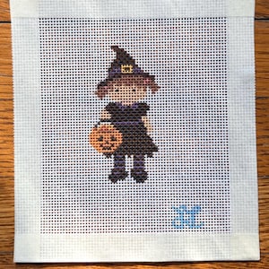 Puede incluir: Un lienzo de punto de aguja con una figura bordada en punto de cruz de un niño disfrazado de bruja, con vestido negro, mallas a rayas moradas y sombrero puntiagudo, sosteniendo un cubo de calabaza naranja, con iniciales azules.