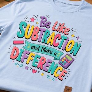 以下が含まれることがあります： 「Be Like SUBTRACTION and Make a DIFFERENCE」という文字が書かれたカラフルなグラフィックが特徴のライトブルーのTシャツ。デザインには、紫色の電卓、マイナス記号、鮮やかな色の星やハートなどの装飾要素が含まれています。