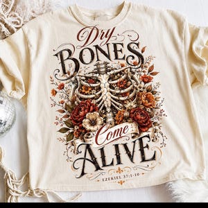 May include: A cream-coloured t-shirt featuring a graphic of a human rib cage intertwined with red, orange, and white flowers. The text reads Dry BONES Come ALIVE and EZEKIEL 37:1-10 in a decorative font. The design uses dark brown and red ink.