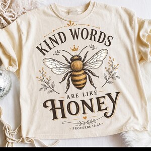 May include: A cream-colored t-shirt featuring a central graphic of a bee with wings spread. Above the bee, the text reads KIND WORDS, and below, ARE LIKE HONEY, followed by PROVERBS 16:24. The design includes small floral sprigs and a crown.