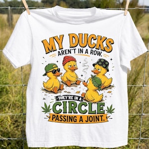May include: A white t-shirt featuring a graphic of four yellow ducks wearing hats and sitting in a circle. The text reads, "MY DUCKS AREN'T IN A ROW. THEY'RE IN A CIRCLE PASSING A JOINT." The design includes small green leaf illustrations.