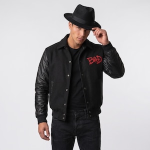 Op de afbeelding: Een zwart varsity-jack met leren mouwen en een body van wolmix, voorzien van het woord BAD geborduurd in rode draad op de borst. Het jack heeft een knoopsluiting, geribbelde boorden en een kraag, gecombineerd met een zwarte fedora-hoed.