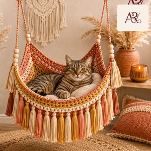 May include: A brown tabby cat resting inside a hanging macrame hammock. The hammock features a woven pattern in shades of orange, yellow, and cream, accented with wooden beads on the suspension ropes and decorative tassels along the bottom edge.