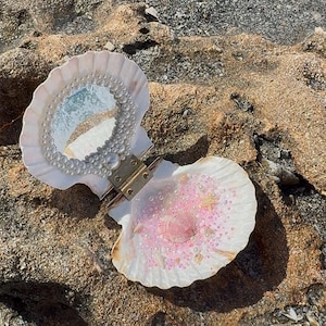 May include: A hinged clam shell compact featuring a circular mirror framed by white pearls on the top interior. The bottom interior is filled with pink sand, small white pearls, and tiny seashells, including a central pink scallop shell.