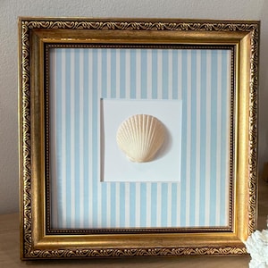 May include: A square wall art piece featuring a single off-white scallop shell centered on a white square mat. The background behind the mat consists of vertical light blue and white stripes. The entire display is enclosed in a gold frame.