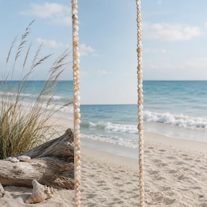 May include: A tall, rectangular floor mirror featuring a frame entirely covered in small, light-colored, rounded seashells. The mirror surface reflects the surrounding beach landscape, showing the sand, ocean waves, and blue sky.