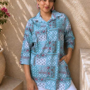 Puede incluir: Una camisa tipo túnica de color azul claro con un patrón de patchwork de diseños florales y geométricos en tonos blanco, rosa y azul oscuro. La camisa tiene cuello, una botonadura parcial frontal, mangas tres cuartos y bolsillos laterales.