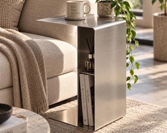 May include: A brushed metal C-shaped side table with a smooth, continuous surface. The table features an integrated middle shelf holding a glass reed diffuser and a bottom shelf containing two books titled The Kinfolk and At Home.