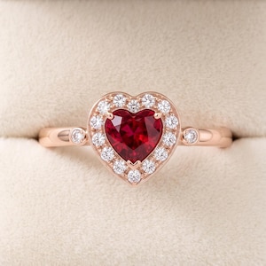 Puede incluir: Un anillo de oro rosa con una piedra preciosa roja central en forma de corazón rodeada por un halo de pequeñas piedras blancas redondas. La banda es lisa y pulida, con una pequeña piedra blanca engastada en el metal a cada lado del centro.
