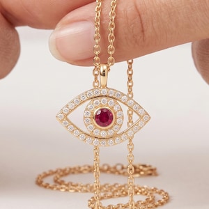 May include: A gold necklace featuring an eye-shaped pendant. The pendant is encrusted with small white stones and has a single round red stone in the center. The piece hangs from a delicate gold chain with a matching metallic finish throughout.