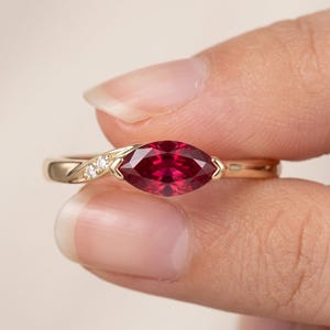 Op de afbeelding: Een gouden ring met een centrale dieprode edelsteen in marquise-slijpvorm. De ringband heeft aan één kant een verfijnd detail, bezet met twee kleine, heldere, rond geslepen witte steentjes die zorgen voor een subtiele schittering.