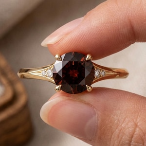 Puede incluir: Un anillo de oro con una gran piedra preciosa granate redonda de color rojo intenso sujeta por cuatro garras. La banda está hecha de oro amarillo pulido e incluye dos pequeñas piedras de acento claras a cada lado del engaste central simétrico.