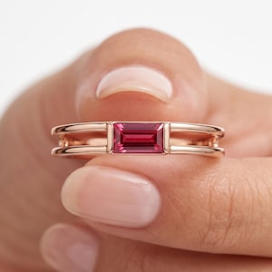 Puede incluir: Un anillo de oro rosa con una sola piedra preciosa rosa de corte baguette rectangular engastada horizontalmente en el centro. La banda está diseñada con un estilo de vástago dividido, creando dos líneas paralelas que se unen en el engaste.