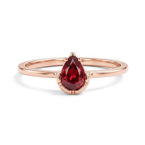 May include: A rose gold ring featuring a single pear-shaped deep red gemstone set in a prong setting. The band is thin and smooth, showcasing a polished metallic finish that complements the vibrant, faceted red stone at the centre of the piece.