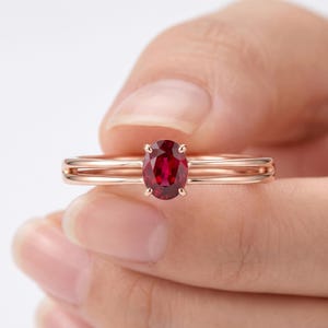 May include: A rose gold ring featuring a single oval-cut deep red ruby gemstone set in a four-prong setting. The band is constructed with two parallel thin metal wires that meet at the center to hold the faceted stone securely in place.