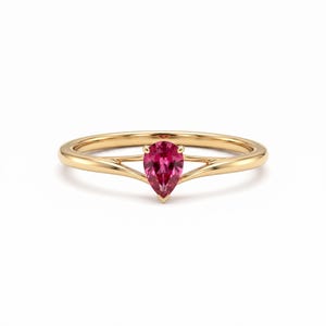 Puede incluir: Un anillo de oro con una piedra preciosa de color rosa intenso en forma de pera, montada en un engaste de garras. La banda es lisa y pulida, con un diseño de vástago dividido que se estrecha al llegar a la piedra central, creando un aspecto limpio.