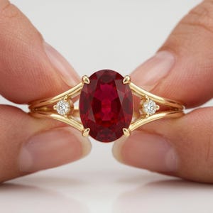 May include: A yellow gold ring featuring a large, oval-cut, deep red ruby as the centre stone. The band splits into two delicate arms on each side, with a small, round, brilliant-cut white diamond set into each side of the gold shank.