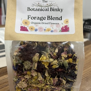 Op de afbeelding: Een doorzichtige plastic zak met een bruine papieren bovenkant gevuld met een mix van gedroogde bloemen. Het etiket vermeldt The Botanical Binky Forage Blend Organic Dried Flowers en toont illustraties van gele, roze en blauwe bloemen.