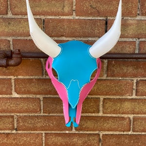 May include: A decorative cow skull with a vibrant color scheme. The skull features a turquoise center, pink accents, and white horns. The edges are stitched with white thread, creating a striking contrast against the colorful design. The skull is mounted on a brick wall.