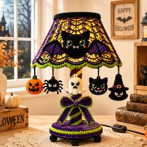 May include: A crocheted table lamp featuring a purple and green spiderweb shade topped with a black bat. The base is wrapped in purple and green yarn with a bow. Hanging charms include a pumpkin, spider, ghost, cat, and witch hat.