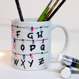 Può includere: Tazza in ceramica bianca con lettere nere che compongono l'alfabeto, decorata con luci colorate. La tazza contiene pennelli e un tubetto di vernice verde. Un oggetto creativo e artistico.
