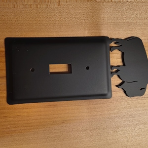 May include: A matte black rectangular light switch cover plate featuring a silhouette of a buffalo extending from the right side. The plate includes a central rectangular cutout for a toggle switch and two circular screw holes for installation.